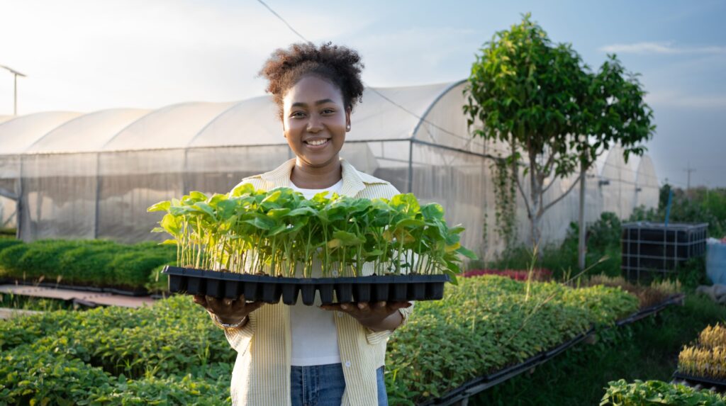 Photograph of young African American woman smiling and holding a tray of vegetables while standing in front of a greenhouse.