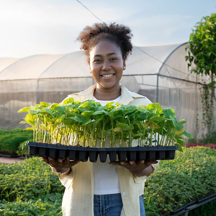 Woman smiling, outside of greenhouse, holding a tray of seedlings