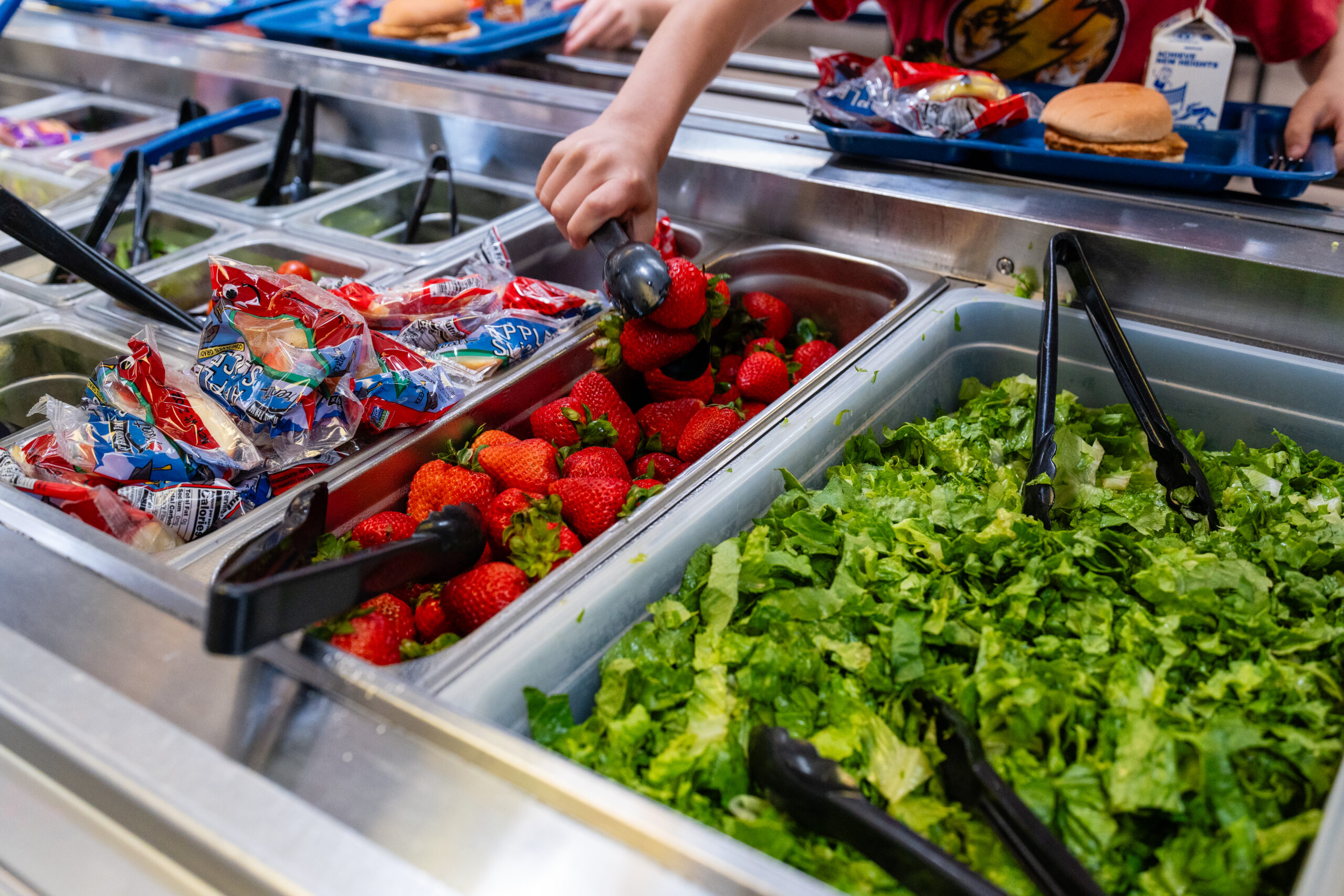 Image of school cafeteria with children's hands taking fruit from salad bar.